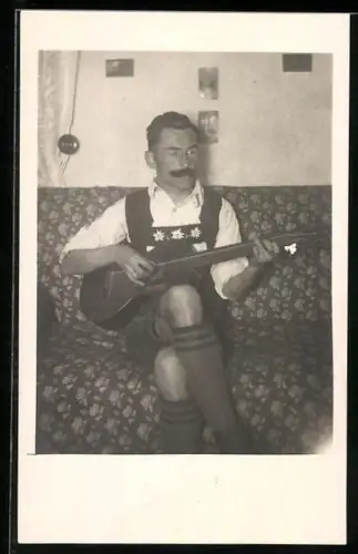 AK Mann in Tracht sitzt mit seiner Gitarre auf einem Sofa