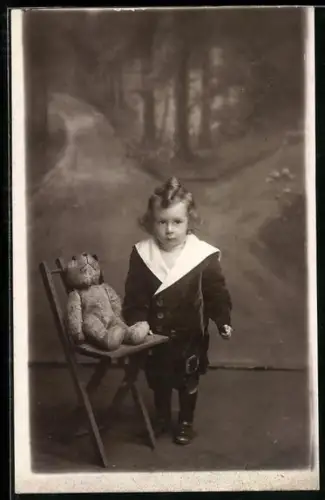 Foto-AK Kleines Kind steht neben einem Teddy auf einem Stuhl