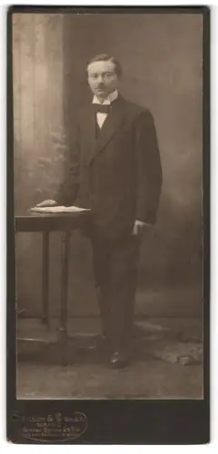 Fotografie Samson & Co., Mainz, Grosse Bleiche 28, Eleganter Herr im Anzug neben Tisch