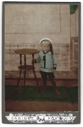 Fotografie unbekannter Fotograf und Ort, Kleiner Junge in Matrosenanzug neben Tisch