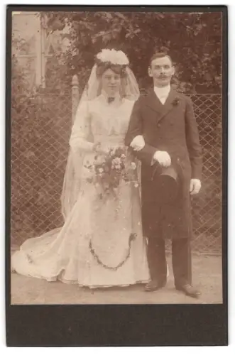 Fotografie unbekannter Fotograf und Ort, Hochzeitspaar mit Blumenstrauss vor einem Zaun stehend