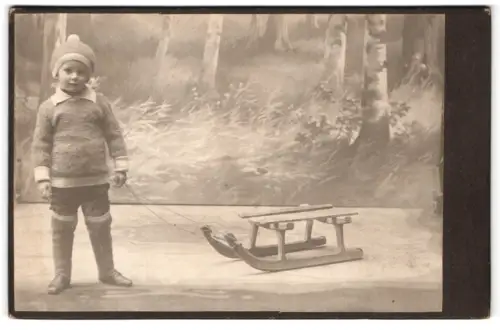 Fotografie unbekannter Fotograf und Ort, Kleiner Junge mit Schlitten vor einer Wald Studiokulisse