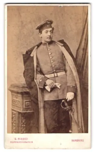 Fotografie E. Bieber, Hamburg, Neuer Jugfernstieg 20, Einjährig-Freiwilliger in Uniform mit Mantel, Krätzchen und Säbel