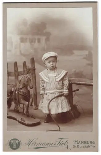 Fotografie Hermann Tietz, Hamburg, Gr. Burstah 12/14, Kleinkind in Matrosenkleid mit Spielzeugpferd und Peitsche