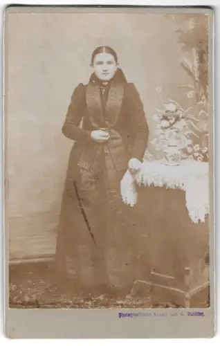 Fotografie Carl E. Schitter, Ort unbekannt, Frau in Tracht neben einem Tisch mit Blumenvase