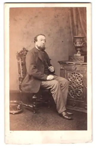 Fotografie unbekannter Fotograf und Ort, Herr sitzt auf Stuhl neben dekorativem Tisch