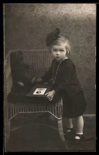 Foto-AK Junges Kind mit Schleife im Haar steht neben Teddy mit einem Fotoalbum