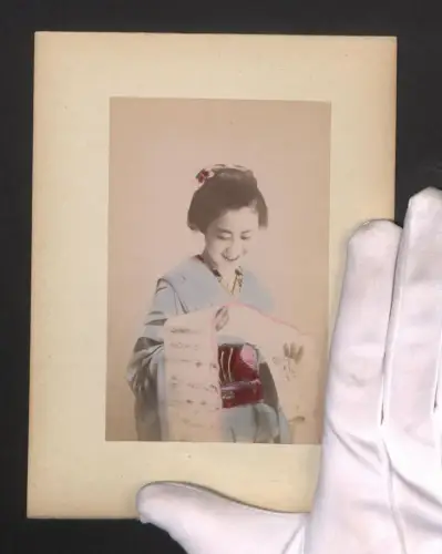 Fotografie Lächelnde Geisha im Kimono liest eine Schriftrolle, Teilansicht von Nagasaki am Ufer, handkoloriert