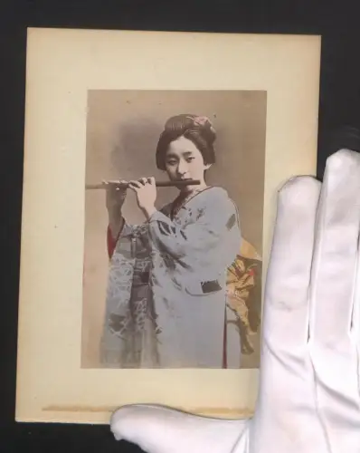 Fotografie Hübsche Geisha im Kimono mit Flöte, Ortsansicht mit Flagge an einer Hütte, handkoloriert