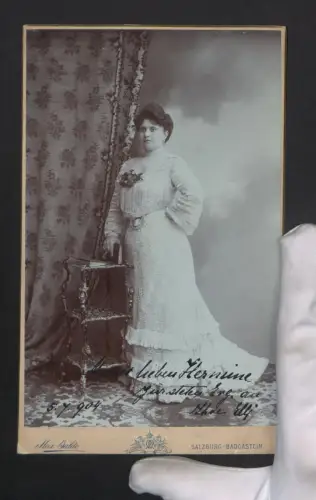 Fotografie Max Balde, Salzburg, Elegante Dame Thea Elli im Kleid neben einem Tisch, 1904