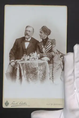 Fotografie Friedr. Schiller, Wien, Mariahilferstr. 107, Portrait eines eleganten Paares an einem Tisch mit Blumen