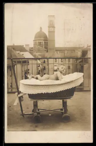 Foto-AK Nacktes Baby bäuchlings im Kinderwagen auf einem Balkon, Turm- u. Kuppelgebäude