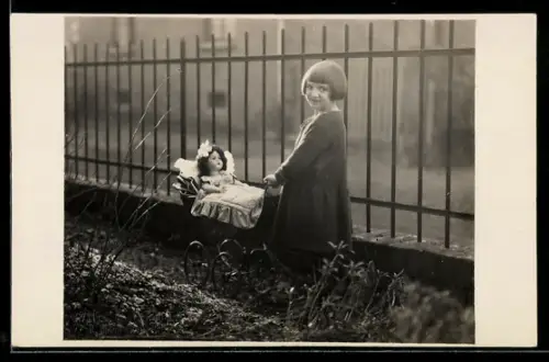 Foto-AK Kleines Mädchen mit Puppe im Puppenwagen neben einem Gartenzaun, 1925