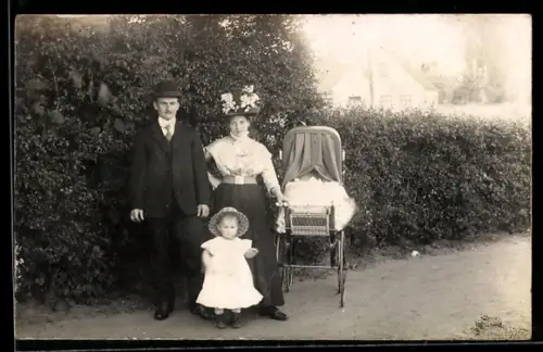 Foto-AK Junges Paar und Kleinkind im Sonntagsstaat vor einer Hecke, Kinderwagen