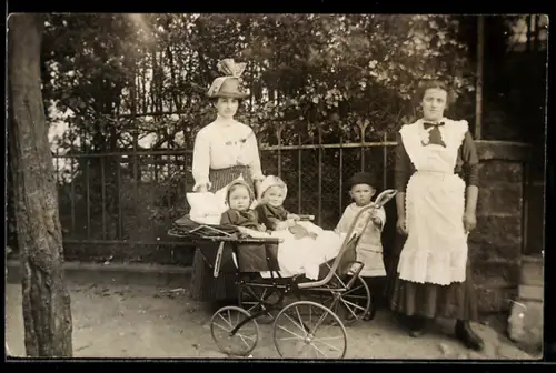 Foto-AK Dame und Dienstmädchen, Knabe u. zwei Kleinkinder im Kinderwagen neben einem Gartenzaun