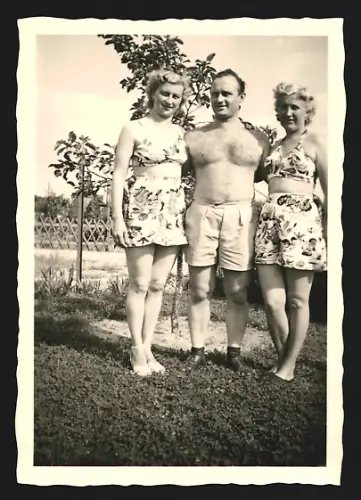 Fotografie Mann steht mit zwei Frauen im Bikini auf einer Wiese