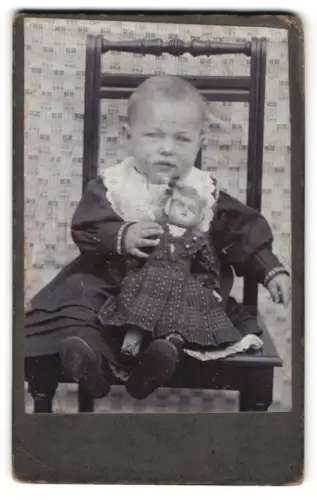 Fotografie unbekannter Fotograf und Ort, Kleinkind mit Puppe auf einem Stuhl sitzend