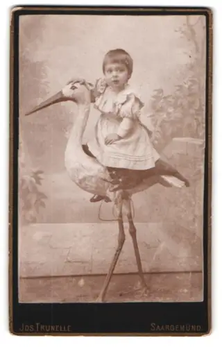 Fotografie Jos. Trunelle, Saargemünd, Kasernstr. 3, Kleinkind auf einer Storchen-Figur