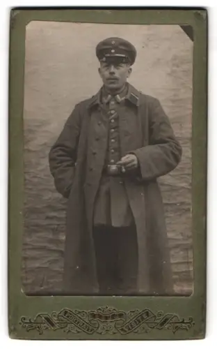 Fotografie unbekannter Fotograf und Ort, Unteroffzier in Feldgrau Uniform mit Mantel, Schirmmütze und Ordensband