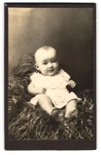 Fotografie unbekannter Fotograf und Ort, Portrait niedliches Kleinkind im weissen Kleidchen auf Fell sitzend
