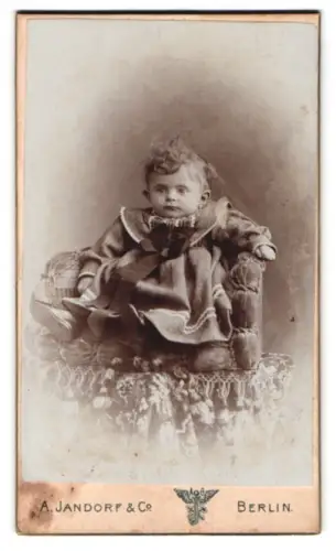 Fotografie A. Jandorf & Co., Berlin, Grosse Frankfurterstr. 113, Kleinkind im Kleid auf einem Sessel