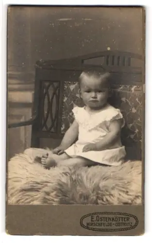 Fotografie E. Ostenkötter, Hirschfelde-Ostritz, Portrait niedliches Kleinkind im weissen Hemd auf Fell sitzend