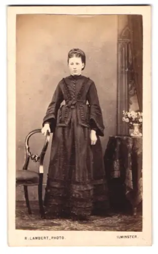 Fotografie R. Lambert, Ilminster, Junge Frau im eleganten Kleid neben Stuhl