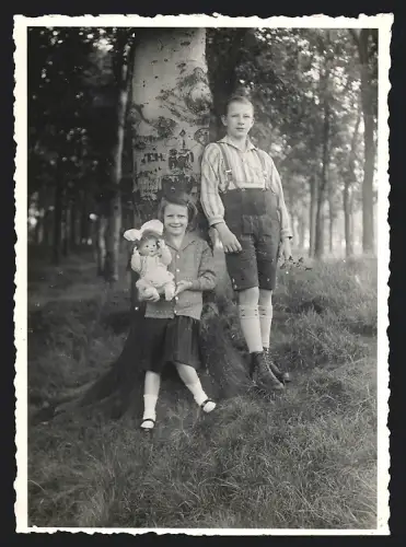 Fotografie Bursche und Mädchen mit Puppe an Buche lehnend