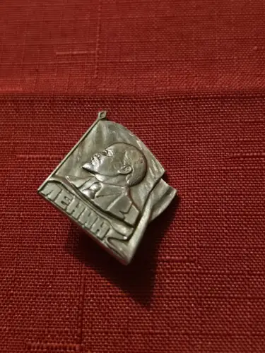 Anstecknadel - Reliefporträt von Wladimir Lenin vor einer wehenden Fahne.